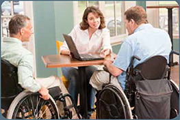 Two disabled veterans in wheelchairs in an office setting working with a woman on a laptop.